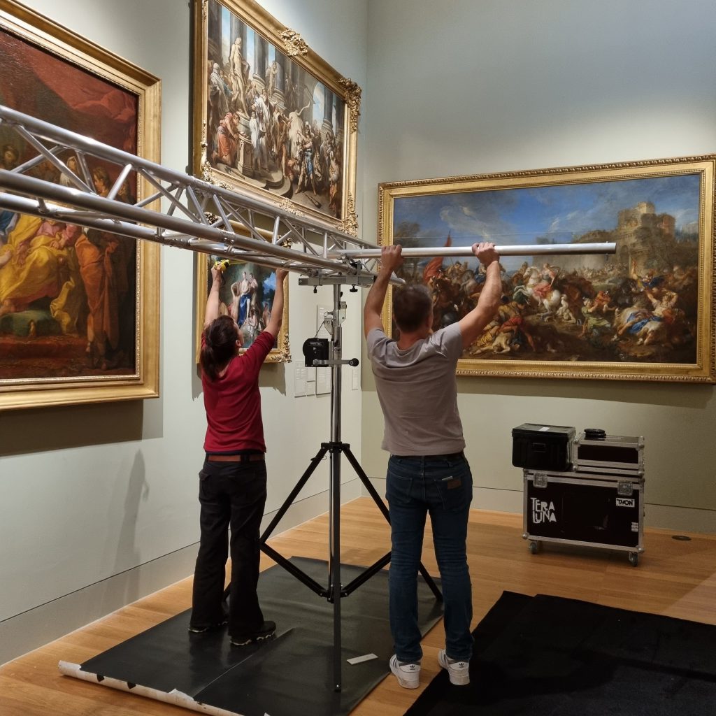 Deux personnes lèvent à bout de bras une barre de structure sur son pied, dans une salle du musée garnie de grands tableaux à cadres dorés.