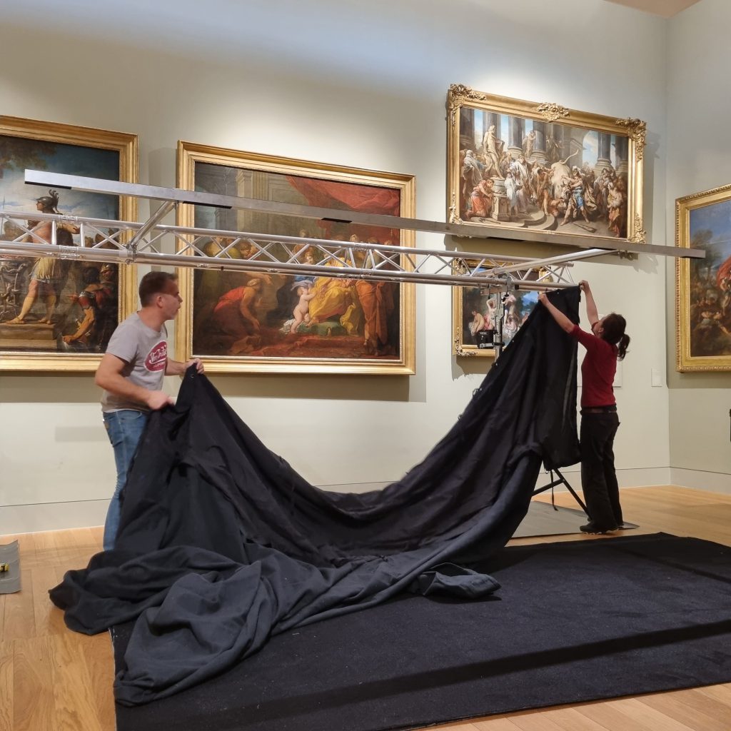 Deux personnes accrochent une grande tenture noire à la structure montée devant le mur de tableaux ; le tissu retombe en plis jusqu’au tapis de sol.