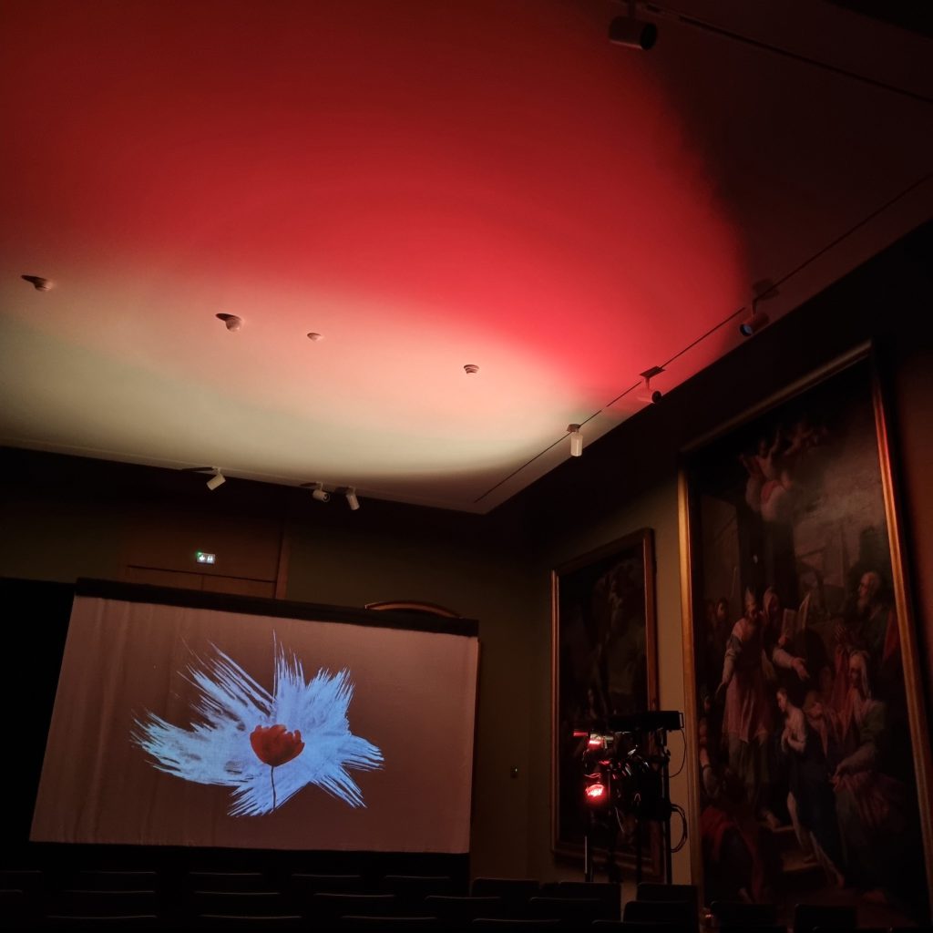 Sur l’écran dressé dans la salle du musée, un coquelicot rouge au cœur d’une gerbe de peinture blanche ; le plafond est noyé de rouge, les chaises encore vides.