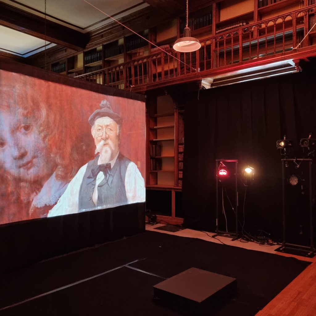 Dans une bibliothèque à galeries de bois, l’écran porte le portrait peint d’un vieil homme à béret et lorgnon ; à droite, la tenture noire et les projecteurs sur pied, la salle encore vide.
