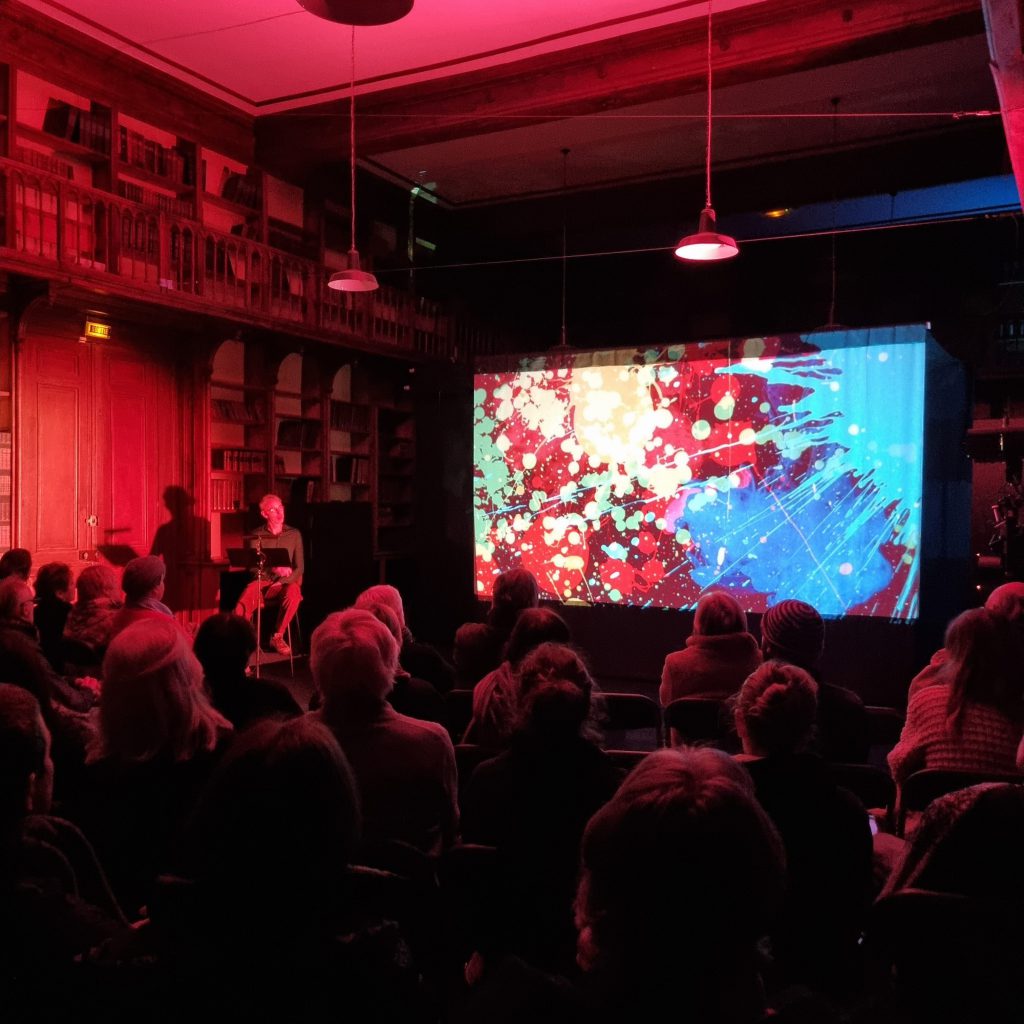 Salle pleine dans la bibliothèque baignée de rouge : le comédien, assis à son pupitre sur la gauche, dit le texte devant une projection de peinture éclaboussée de rouge et de bleu.