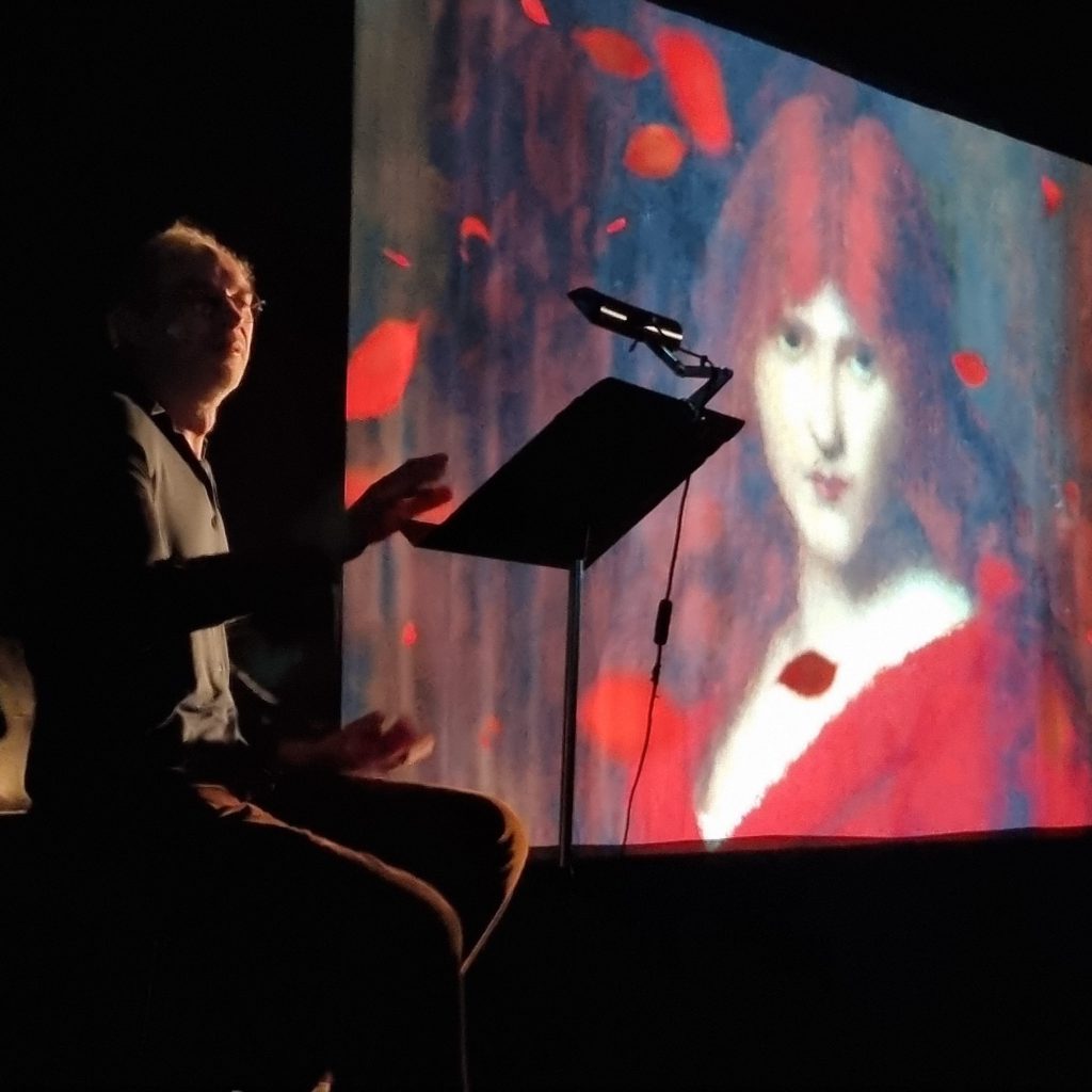 Le comédien, assis à son pupitre et la main levée, devant le portrait projeté d’une jeune femme rousse vêtue de rouge, parmi des pétales en suspension.