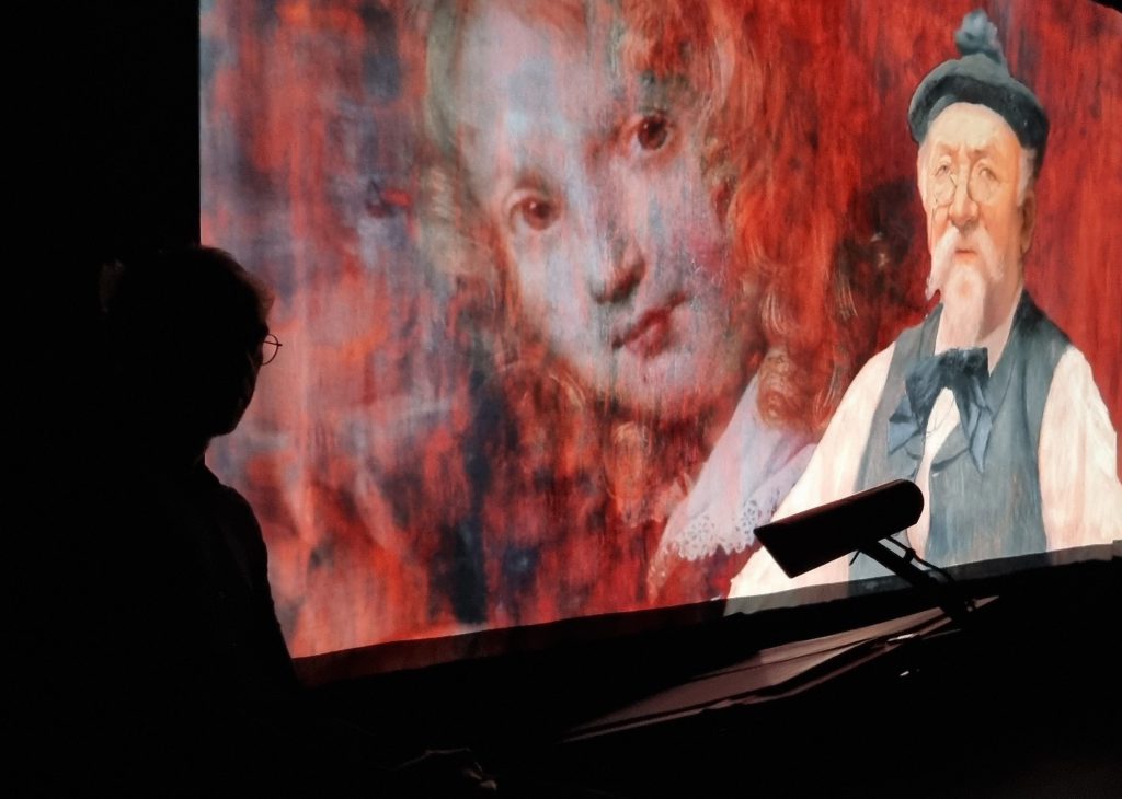 En ombre au premier plan, le profil du comédien à son pupitre ; derrière lui, deux portraits peints se superposent sur l’écran, un visage d’enfant dans la brume rouge et le vieil homme à béret et lorgnon.
