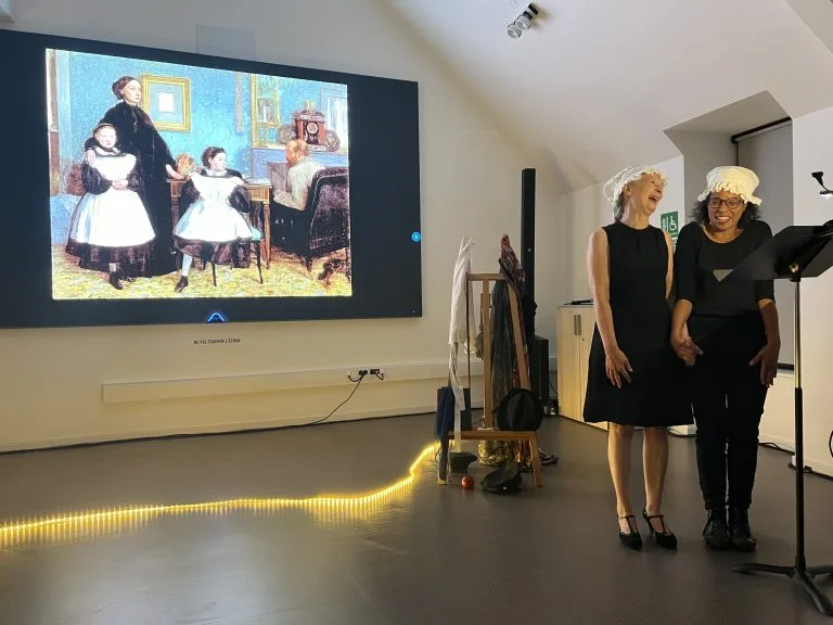 Deux comédiennes en coiffes blanches rient devant l’écran où est projeté un tableau ancien.