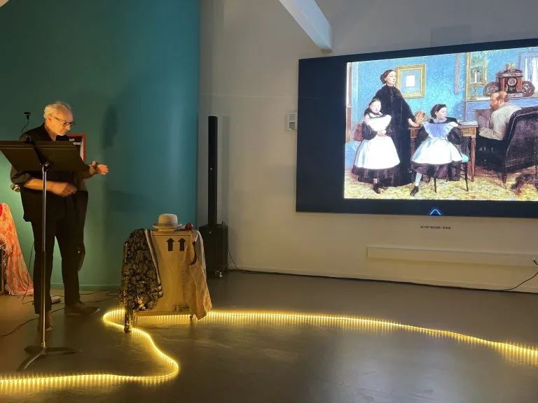 Un comédien lit à son pupitre à gauche ; le tableau projeté occupe toute la droite de la salle.