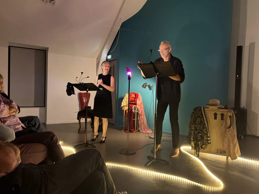 Une comédienne et un comédien à leurs pupitres, une lampe magenta entre eux, le public au premier plan.