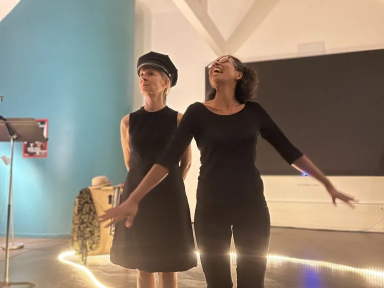 Deux comédiennes en noir, bras ouverts, l’une coiffée d’une casquette.