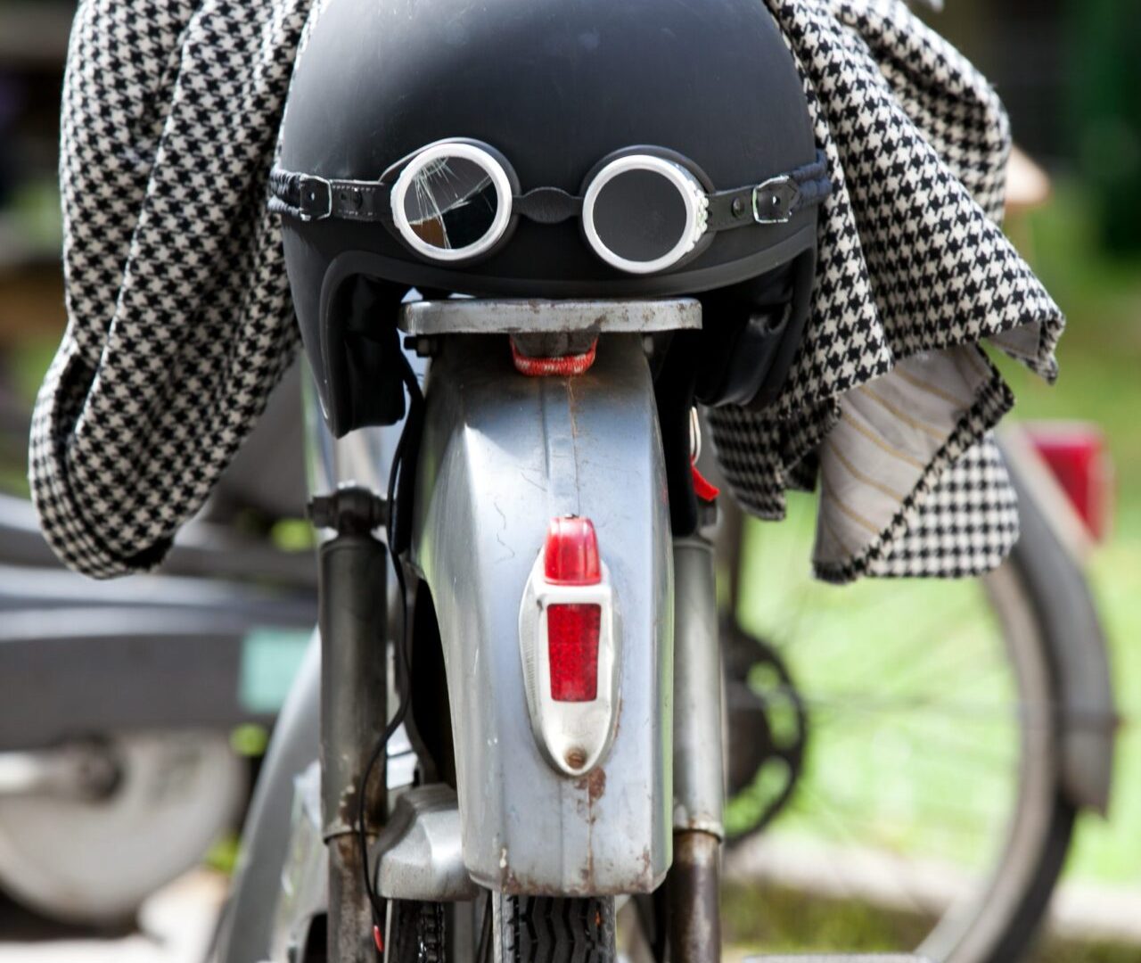 Un casque noir coiffé de lunettes d’aviateur, posé sur le garde-boue d’une mobylette, une veste pied-de-poule jetée par-dessus.
