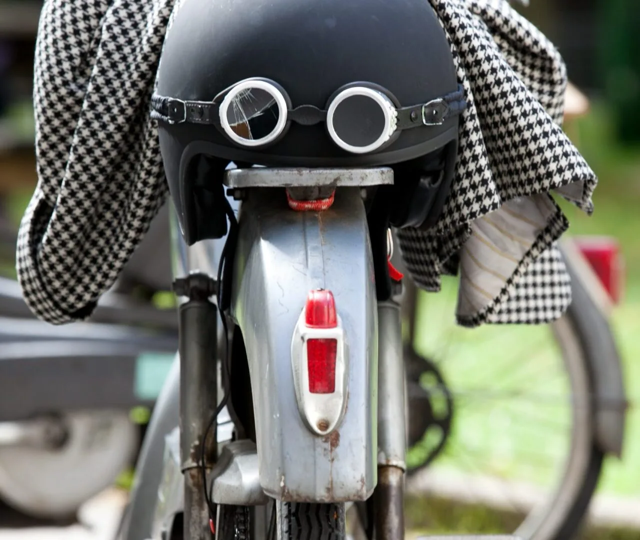 Un casque noir coiffé de lunettes d’aviateur, posé sur le garde-boue d’une mobylette, une veste pied-de-poule jetée par-dessus.