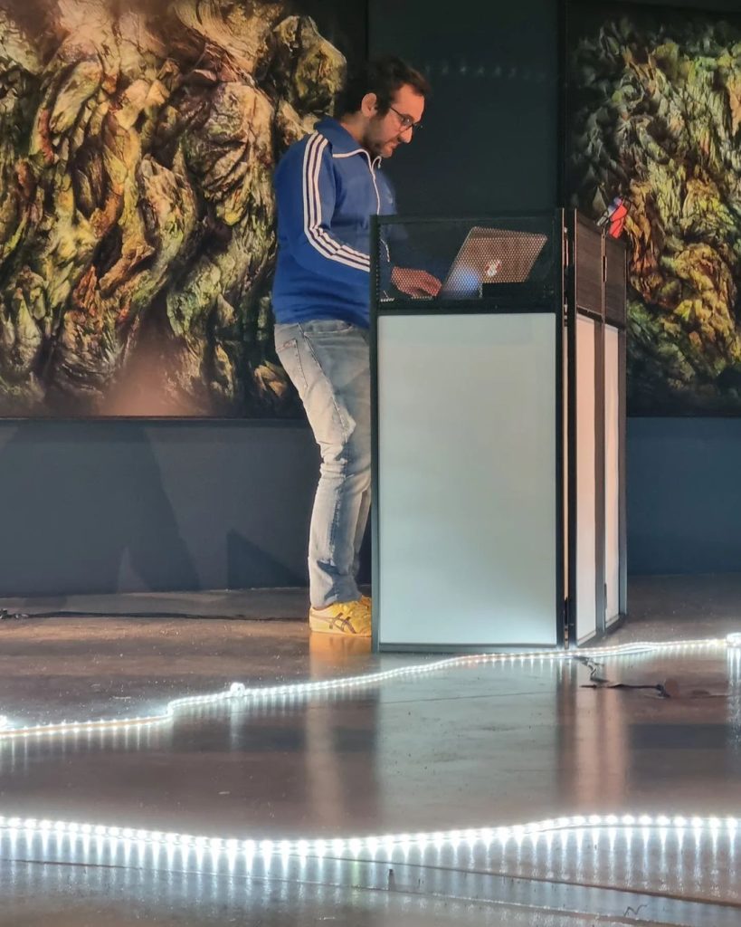 Un musicien en veste de survêtement bleue manipule son ordinateur derrière un pupitre translucide ; au sol, un ruban lumineux serpente devant de grandes photographies d’écorces.